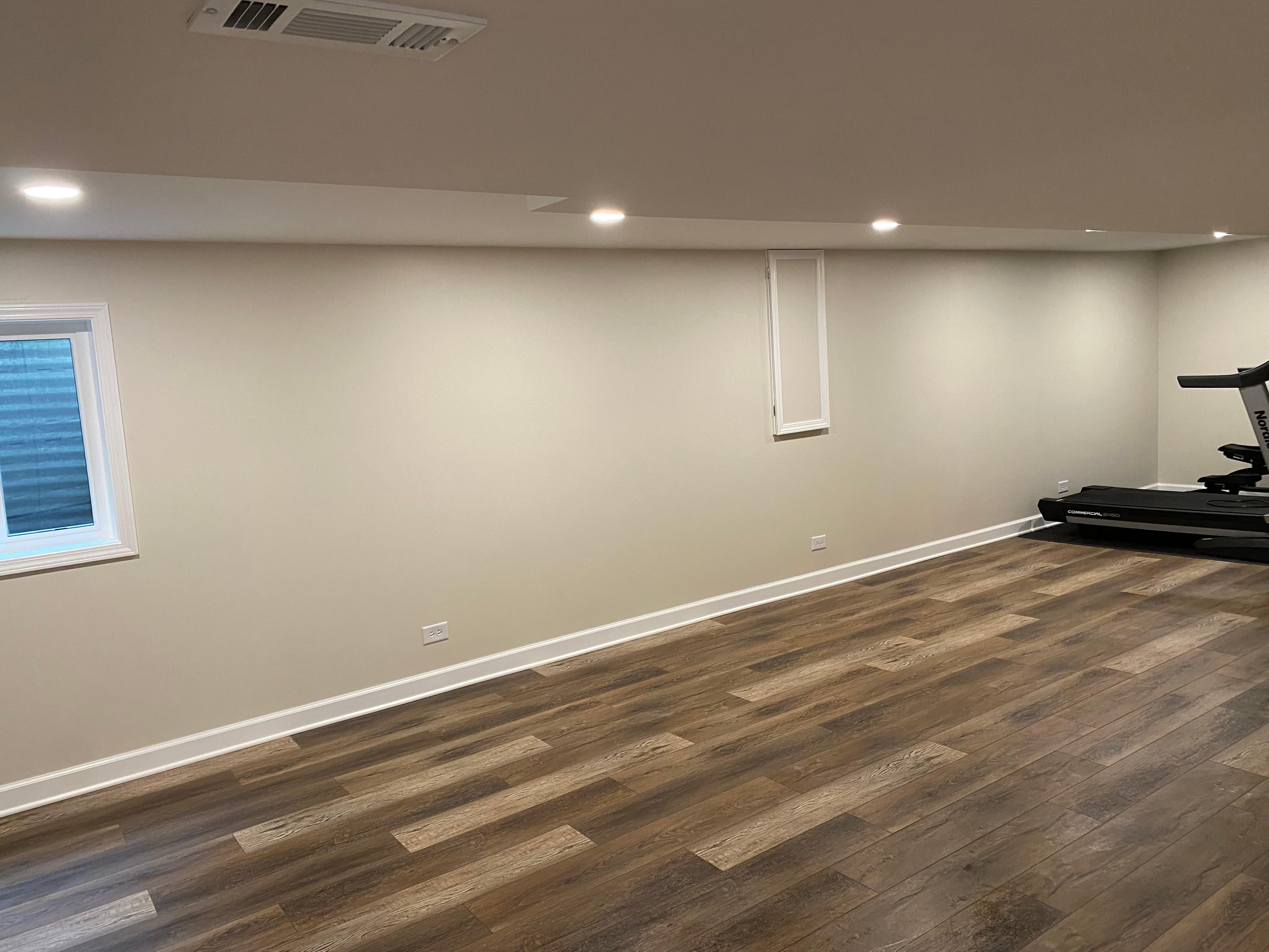 Finished basement with pale walls, wood-look flooring and recessed ceiling lights.